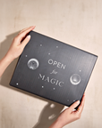 Two hands hold a black gift box emblazoned with white text that reads "Open for Magic" against a beige background. Decorated with tiny stars and circular designs, the box exudes enchantment, suggesting organic botanicals are inside to elevate your wellness routine with the Monthly Magic® First Sips Tea Subscription - Just the Tea!