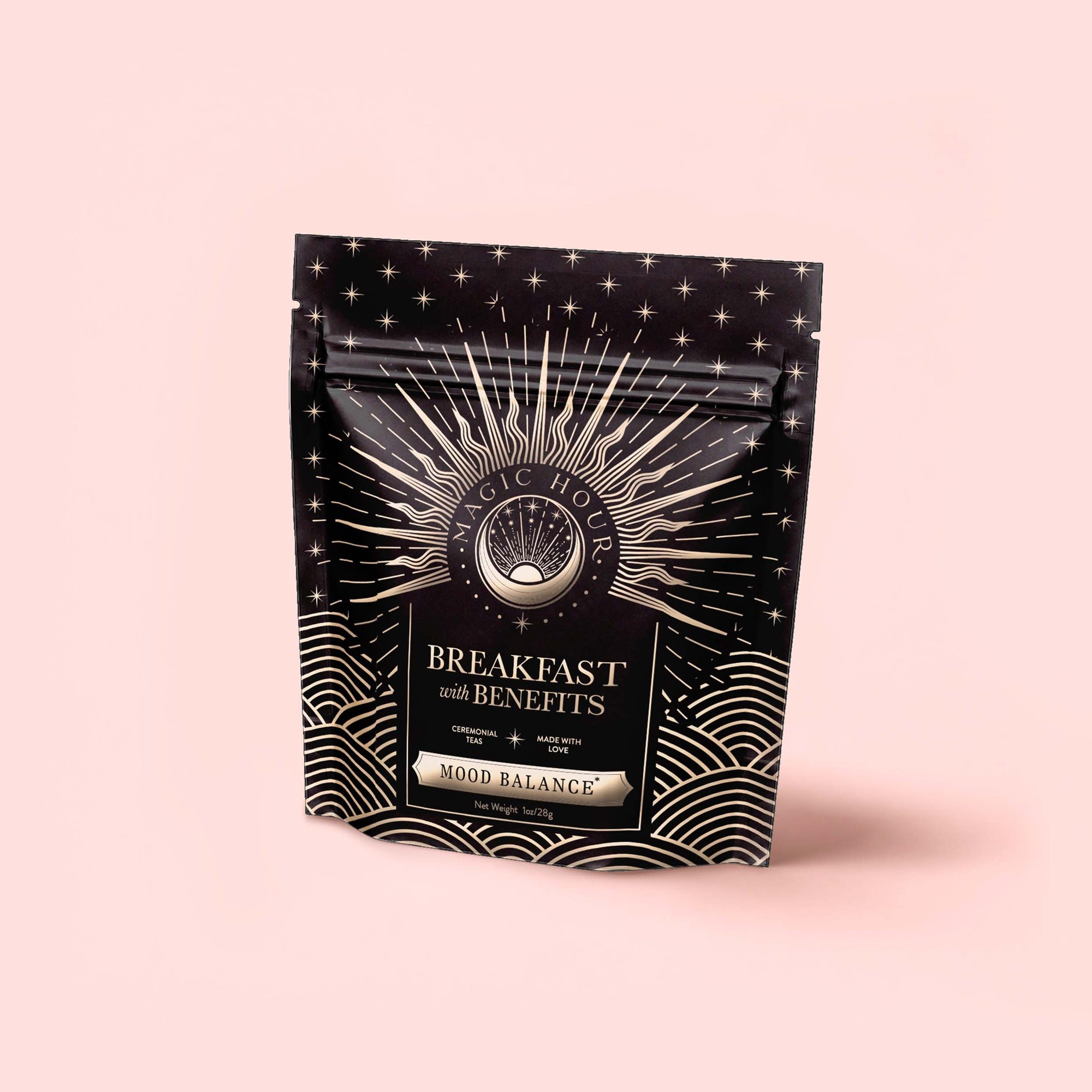 A black and gold resealable pouch labeled "Mood Balance: Breakfast with Benefits" stands upright against a pale pink background, featuring a sunburst and wave design—ideal for enjoying herbal breakfast tea with ashwagandha.