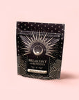 A black and gold resealable pouch labeled "Mood Balance: Breakfast with Benefits" stands upright against a pale pink background, featuring a sunburst and wave design—ideal for enjoying herbal breakfast tea with ashwagandha.