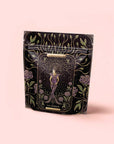 A black resealable bag labeled "The Empress: Lavender Currant Shatavari Cocoa Tea of Nurturing Creativity" features ornate, mystical art of a crowned woman amid roses, with gold, purple, and green on pale pink—perfect for organic herbal tea.