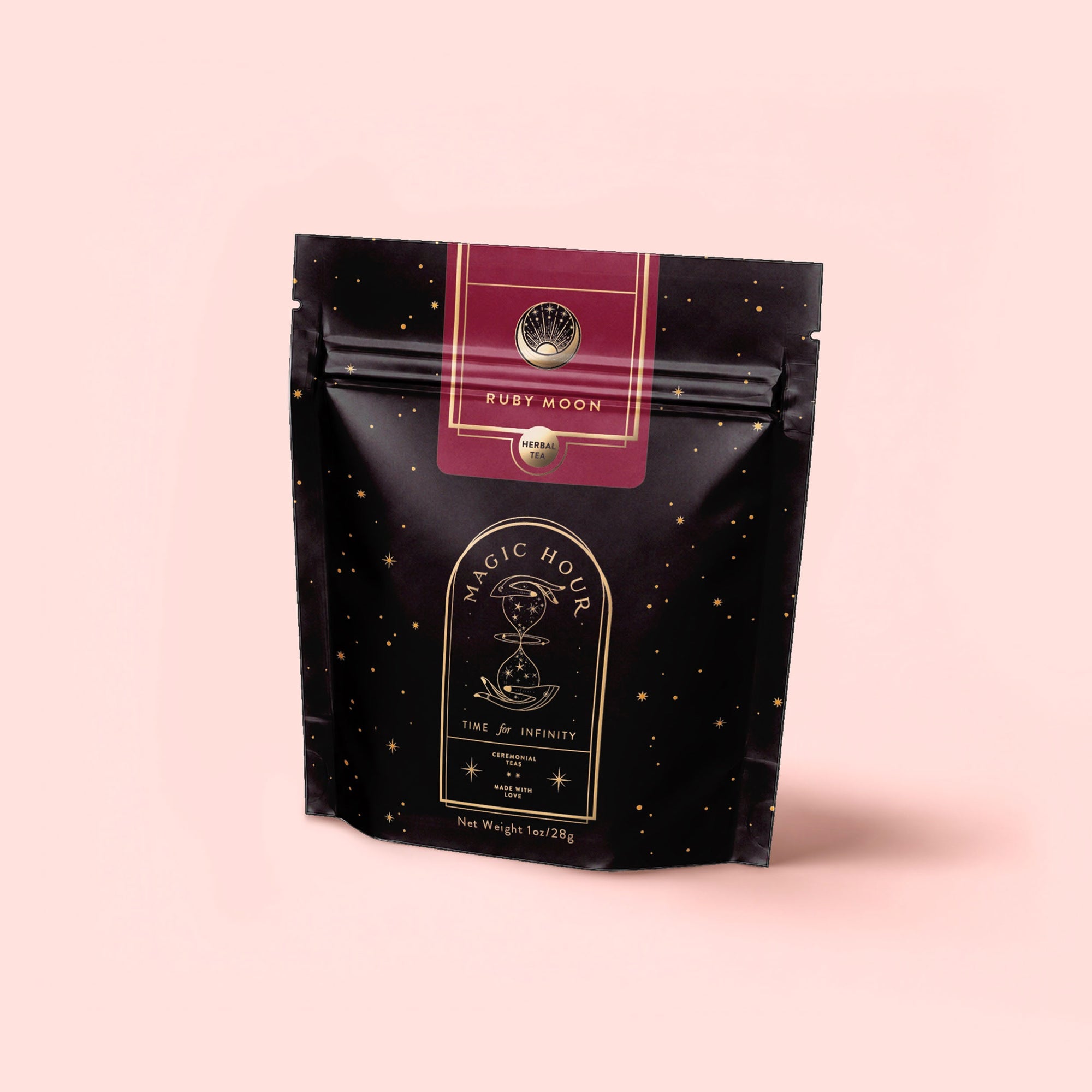 A black and gold speckled resealable pouch labeled "Magic Hour" and "Ruby Moon," from the Herbal Tea Sampler Tea Box: Caffeine-Free Teas for Sleep, Digestion &amp; Immunity, stands upright on a pale pink background with celestial designs.