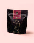 A black and gold speckled resealable pouch labeled "Magic Hour" and "Ruby Moon," from the Herbal Tea Sampler Tea Box: Caffeine-Free Teas for Sleep, Digestion & Immunity, stands upright on a pale pink background with celestial designs.