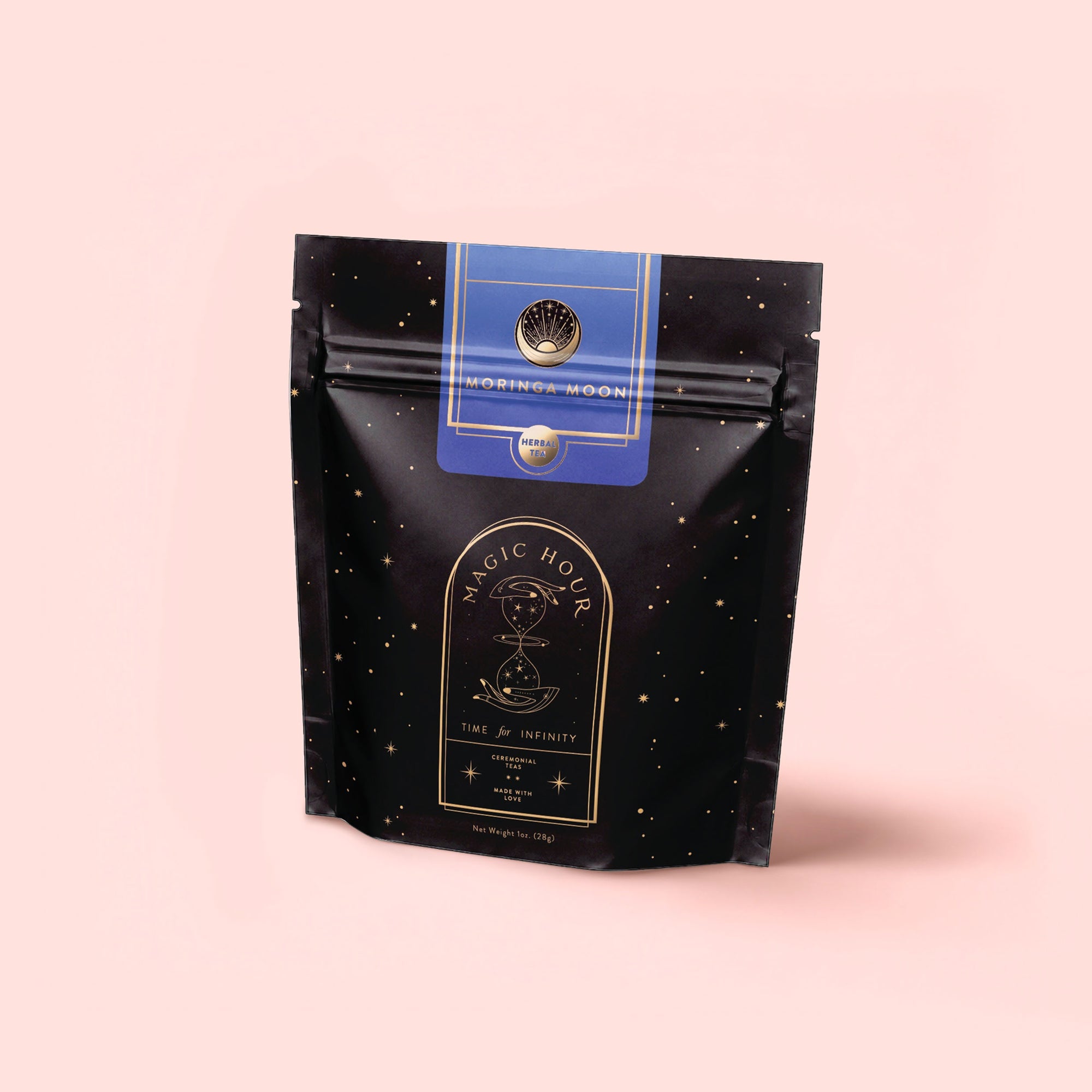 A black and gold resealable package of Magic Hour caffeine-free tea, labeled “Moringa Moon Herbal Tea,” stands upright against a soft pink background with celestial designs and elegant typography.