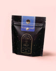 A black and gold resealable package of Magic Hour caffeine-free tea, labeled “Moringa Moon Herbal Tea,” stands upright against a soft pink background with celestial designs and elegant typography.