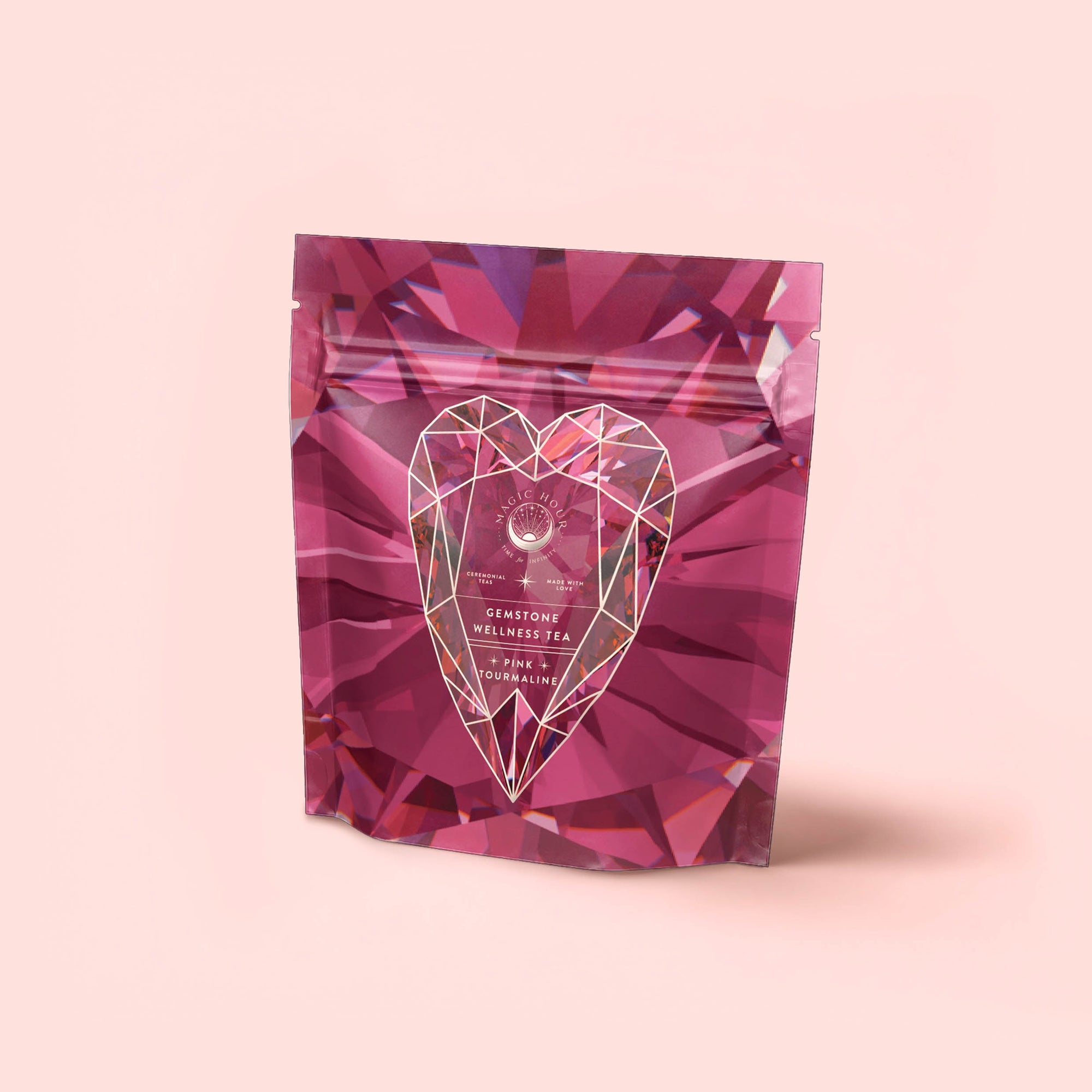 A pink, gem-patterned stand-up pouch with a geometric heart and white text reading "Pink Tourmaline: Organic Dragon fruit Hibiscus White Tea for Women's Health" on the front, set against a pale pink background.