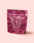 A pink, gem-patterned stand-up pouch with a geometric heart and white text reading "Pink Tourmaline: Organic Dragon fruit Hibiscus White Tea for Women's Health" on the front, set against a pale pink background.