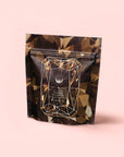A resealable pouch with a geometric gemstone design and dark brown tones, labeled "Black Onyx - Chaga Maca Toffee Elixir for Energy, Immunity & Good Fortune," features a caffeine-free blend against a pale pink background.