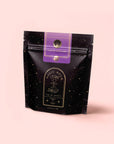 A black, star-speckled "Magic Hour" pouch with gold accents and a "Lucid Dreams" tag displays caffeine-free herbal tea against a pink background—an ideal pick for the Herbal Tea Sampler Tea Box: Caffeine-Free Teas for Sleep, Digestion & Immunity.