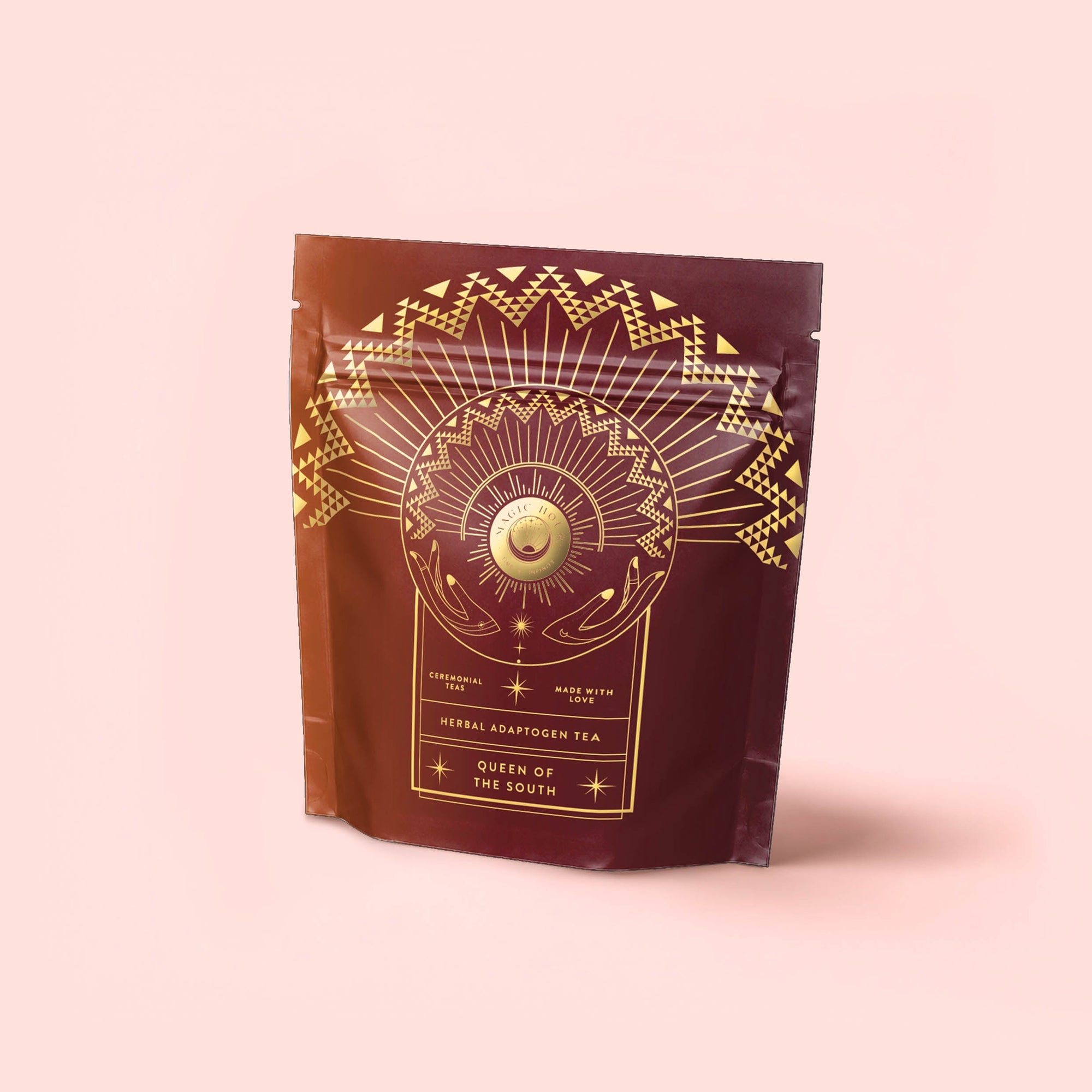 The Queen of the South Cocoa Detox Tea pouch is maroon with gold geometric and celestial designs—rays, zigzags, and a central circle—all set against a pale pink background, evoking Café de Olla style.