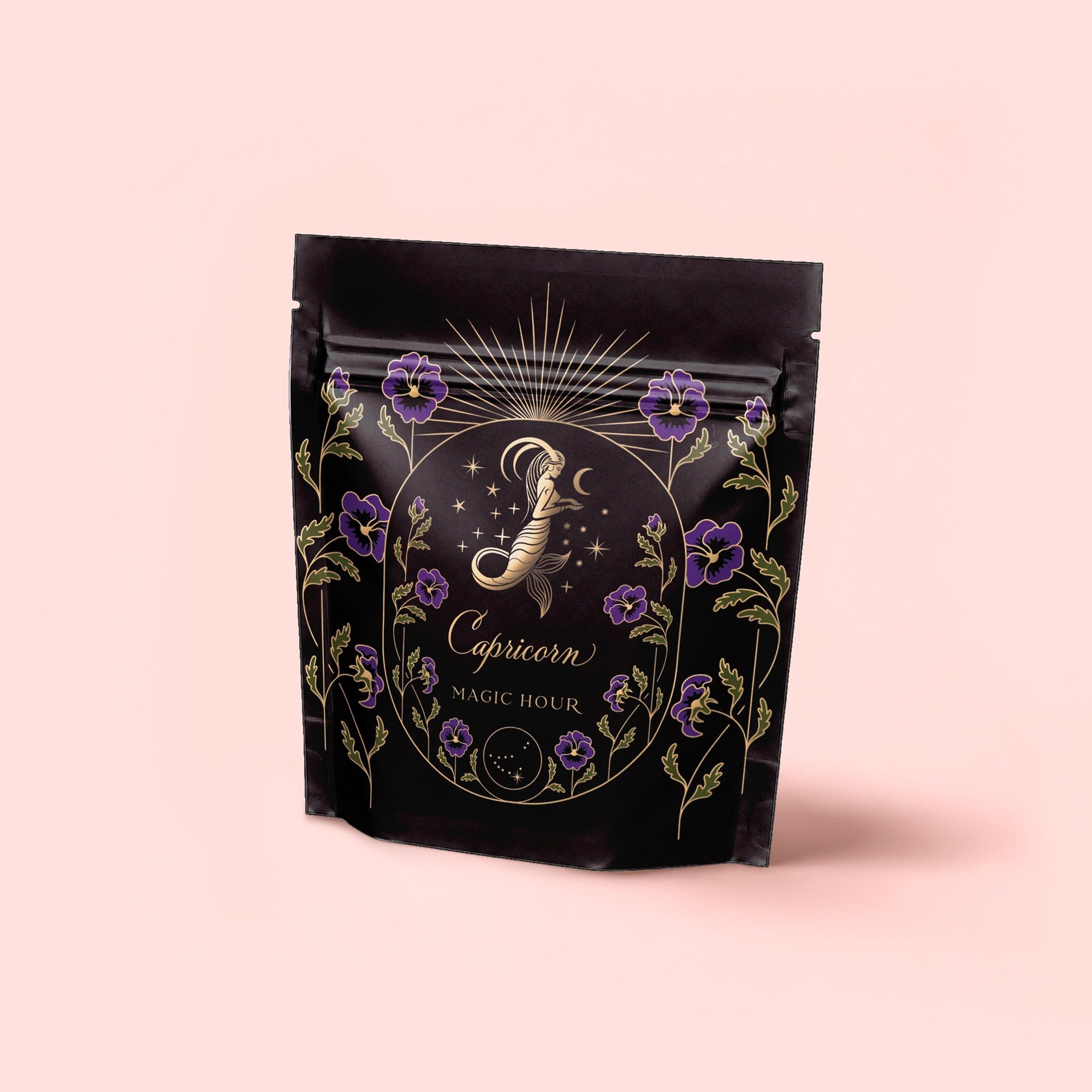 A black resealable bag with gold and purple floral designs, labeled "Capricorn: Maple Oolong Tea" and "Magic Hour," features a crescent moon, stars, and an ornate letter "J"—crafted with metabolism-boosting Se Chung Oolong on a pale pink background.