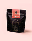 A black resealable pouch labeled "Magic Hour" with gold stars and a celestial design contains Rise & Shine Black Tea, an adaptogenic wellness blend. The pouch features a pink label and stands against a pale pink background.