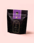 A black and gold resealable pouch labeled "Zhena’s Original Coconut Chai," featuring Fair Trade tea with organic spices, accented in purple on pale pink. The packaging has a starry design and mystical symbols.