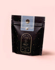 A black pouch labeled "Vanilla Earl Grey: Tea for the Lushness of Life" stands upright against a soft pink background, featuring gold and white text with celestial-themed graphics.