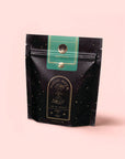 A black resealable pouch labeled "Renewal: Peach-Goji-Rose Oolong Tea" with gold stars and a celestial design stands upright on a pink background. The label features a green top section with a circular emblem, hinting at sweet Peach notes.