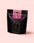 A black resealable tea pouch with star-like dots, labeled "Symbeeosis: Beautifying Immunitea for the Queen Bee," stands on a light pink background, its design hinting at a soothing and beautifying wellness blend inside.