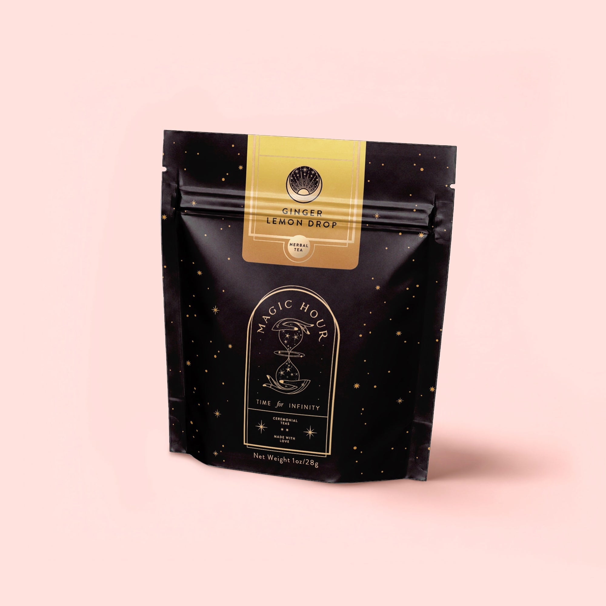 A black, star-speckled resealable pouch labeled "Magic Hour" with a yellow “Ginger Lemon Drop” label stands upright against a pale pink background, highlighting the Ginger Lemon Drop tropical tea blend.