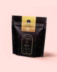 A black, star-speckled resealable pouch labeled "Magic Hour" with a yellow “Ginger Lemon Drop” label stands upright against a pale pink background, highlighting the Ginger Lemon Drop tropical tea blend.