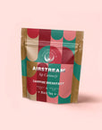A vibrant pouch of Campfire Breakfast: Golden Tipped Assam, Puerh & Lapsang Souchong Pine Smoked Connoisseur's Cup for Nostalgic Mornings stands upright on a light pink background—an ideal smoky tea or coffee alternative.