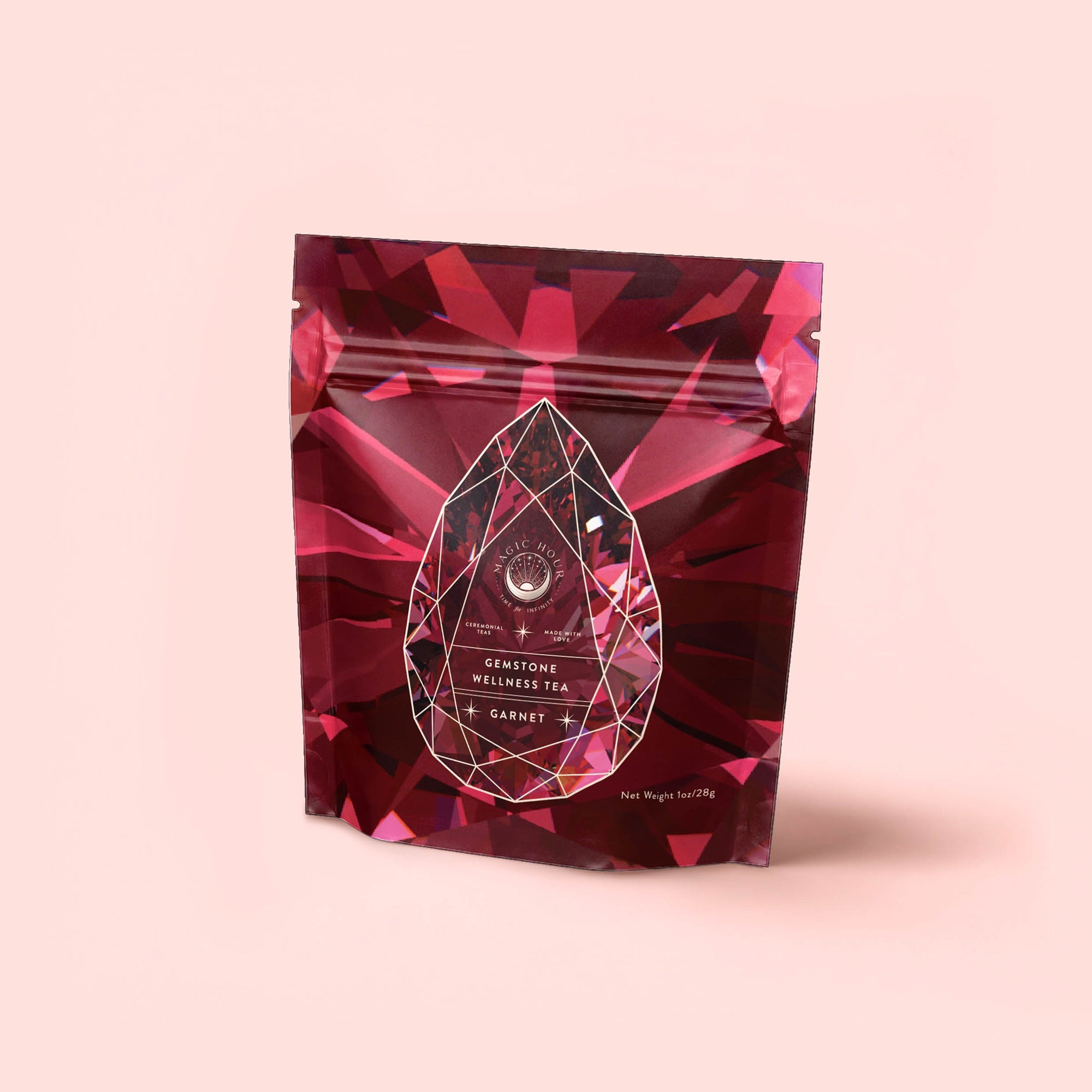 A red, gemstone-patterned resealable pouch labeled "Garnet: Violet Wine Gemstone Wellness Tea" sits against a light pink background, featuring a geometric gem design and white text for this organic herbal tea.