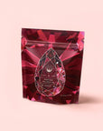 A red, gemstone-patterned resealable pouch labeled "Garnet: Violet Wine Gemstone Wellness Tea" sits against a light pink background, featuring a geometric gem design and white text for this organic herbal tea.