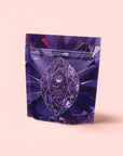 A purple, resealable pouch labeled "Amethyst: Strawberry-Passionfruit Gemstone Wellness Tea" with crystal designs stands upright on a light pink background—ideal for those seeking spiritual clarity with organic herbal tea.