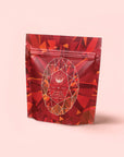 A red and orange resealable pouch with geometric patterns, featuring an oval label that reads “Carnelian: Caffeine-Free Apricot Amaretto Tea” on a light pink background.