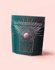 A teal and black resealable pouch labeled "Queen of Wellness: Women's Hormone Balancing Tea for PMS, Healthy Cycles & Menopause" features a pink geometric and celestial design with hands holding a radiant sun on a light pink background.