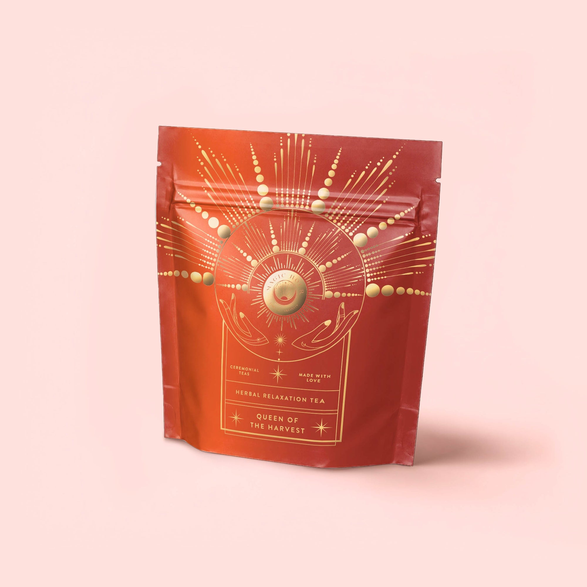 A red resealable pouch with a gold cosmic design—radiating lines, circles, and hands around a sun—on a pale pink background. The label reads: "Queen of the Harvest : Herbal Relaxation Tea. Caffeine-Free.