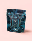 A teal and dark blue resealable pouch of caffeine-free Herbal Tea Sampler Tea Box for sleep, digestion & immunity, with a gemstone-inspired geometric design, is labeled against a soft pink background.