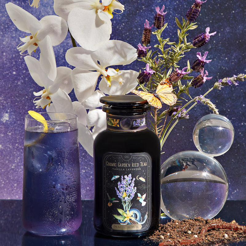 A glass of purple Vanilla Lavender Black Iced Tea with a lemon slice, a Cosmic Garden Iced Teas bottle, white orchids, lavender sprigs, a butterfly, and two glass orbs set against a starry cosmic backdrop.