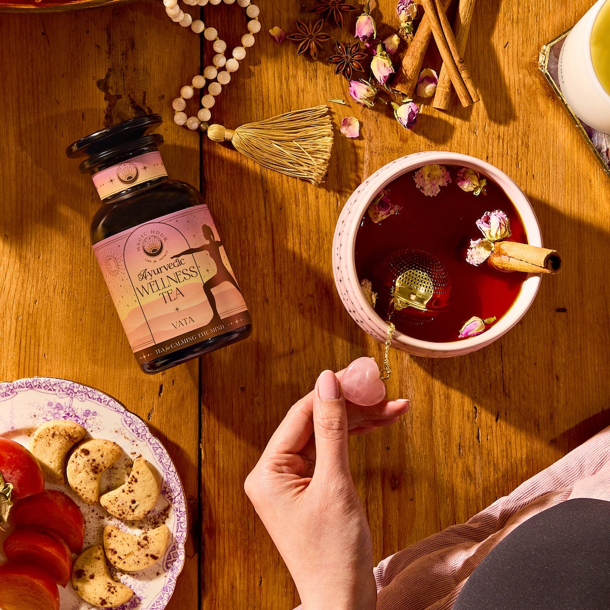 A hand holds a pink crystal by a cup of herbal tea with rose petals and cinnamon. Nearby, a jar labeled "Vata Herbal," sliced cookies, fruit, beads, and a broom rest on a wooden table.