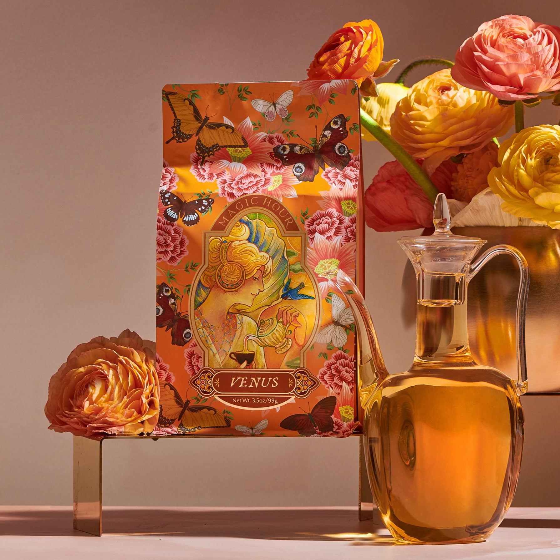 A floral-themed product package labeled "Venus - Tea for Beauty Sleep" with bright, colorful illustrations is displayed alongside a clear glass jug of golden liquid, possibly oil. In the background, a copper vase is filled with vibrant, multicolored ranunculus flowers and botanicals, creating a warm, inviting scene.