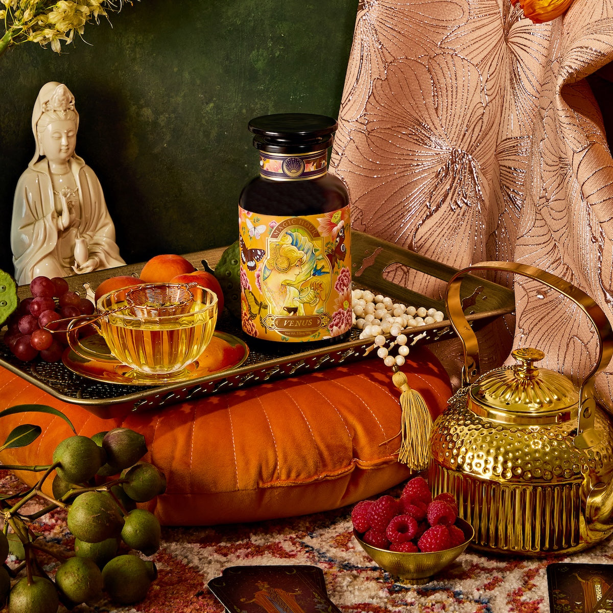 A vibrant display with a golden teapot, glass teacup of Venus - Tea for Beauty Sleep, colorful tea container, fresh grapes and raspberries, small statue, all set against floral pattern fabric.