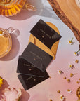 A set of black "Magic Hour" tea packets is arranged on a pink and beige surface with flowers and tea accessories. Nearby, there is a half-full teacup, a glass teapot, yellow and white flowers, scattered chamomile, and a Gift Card for Club Magic Hour to redeem during checkout for the perfect gift of choice.