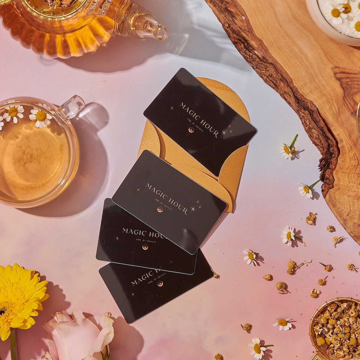 A flat lay of black Gift Card gift cards fanned out on a tan cardholder, surrounded by chamomile flowers, a cup of tea, a teapot, and scattered tea leaves on a wooden board and pastel surface.