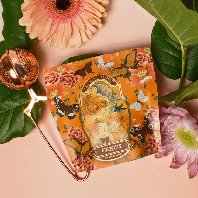 A vibrant tea package named "Venus" from Magic Hour, adorned with Art Nouveau-style illustrations of a woman and child surrounded by roses and butterflies, is part of The Best of Winter Box. This exquisite set boasts over 200 cups of winter astrology teas. The collection is beautifully displayed on a floral background alongside a gold tea infuser and fresh flowers.