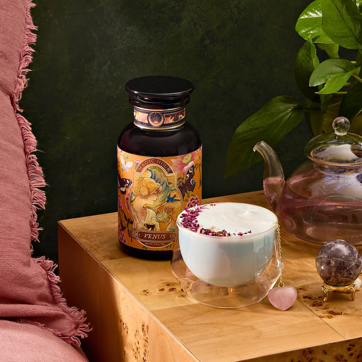 A serene setup showcases Venus - Tea for Beauty Sleep in a jar with a vibrant label next to a double-walled glass cup brimming with frothy, rose petal-adorned, caffeine-free blend. Located on wooden surface, it is accented by pink fabric, crystal decor, and leafy plant for beauty-enhancing hydration.