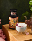 A serene setup showcases Venus - Tea for Beauty Sleep in a jar with a vibrant label next to a double-walled glass cup brimming with frothy, rose petal-adorned, caffeine-free blend. Located on wooden surface, it is accented by pink fabric, crystal decor, and leafy plant for beauty-enhancing hydration.