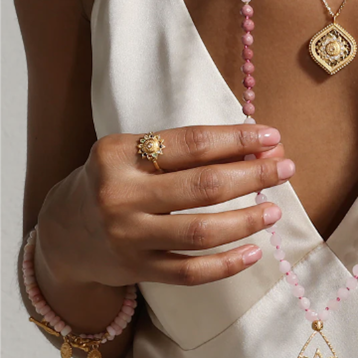 A person wearing a white satin top displays a pink beaded necklace, a gold pendant featuring a sunburst design, and an 18kt gold-plated lotus ring with healing gemstones, along with a matching bracelet. The person's hand, adorned with the Magic Hour Vibrant Self Multi Stone Lotus Ring, delicately holds part of the necklace.