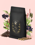 A black tea bag with a gold, celestial "Wings Magic Hour" label sits on pink, surrounded by elderberries, green leaves, licorice roots, and loose organic black tea from the Virgo Tea of Virtue, Wit & Meticulous Magic blend.