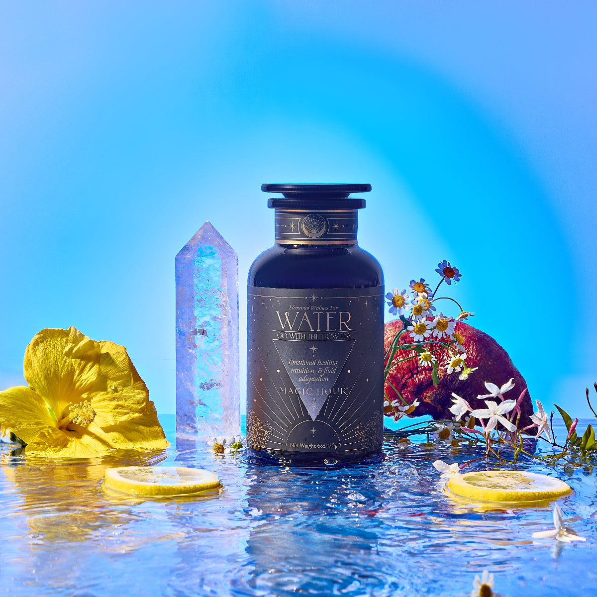 The Water dark glass tea jar stands on reflective water with yellow flowers, lemon slices, white wildflowers, a crystal, chamomile, and pomegranate around it, set against a vibrant blue background.