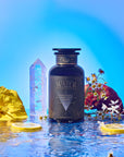 The Water dark glass tea jar stands on reflective water with yellow flowers, lemon slices, white wildflowers, a crystal, chamomile, and pomegranate around it, set against a vibrant blue background.