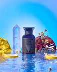 A dark blue Water bottle rests on a reflective watery surface, surrounded by yellow flowers, sliced citrus, white daisies, a crystal, and a halved plum, set against a vivid blue backdrop.
