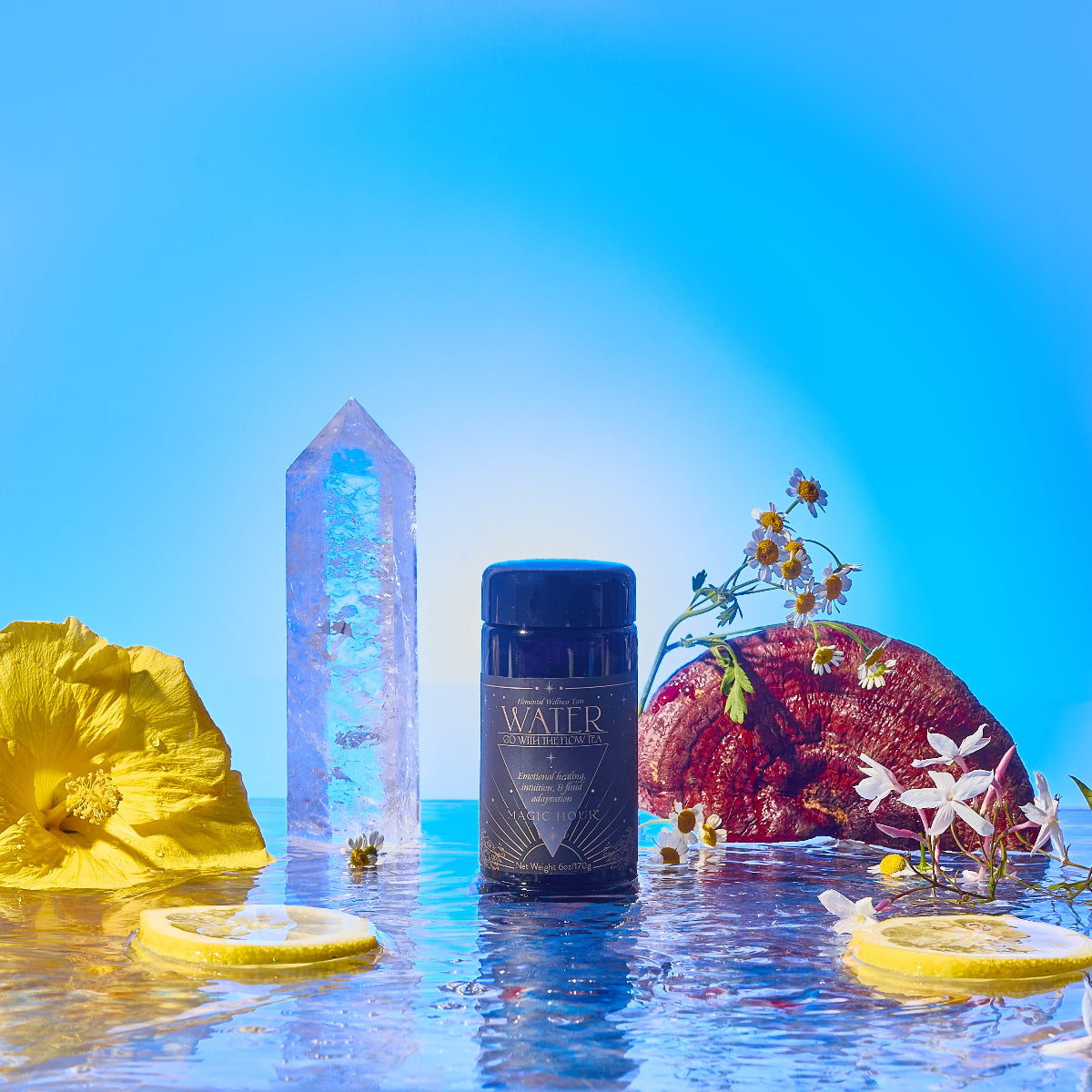 A dark jar labeled "Water" rests on a reflective surface amid a crystal, yellow and white flowers, lemon slices, and a red stone, all set against a bright blue background.