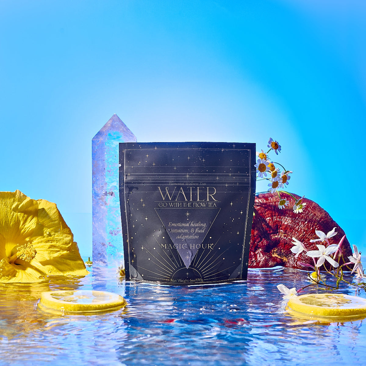 A packet of Water sits on reflective water, surrounded by flowers, lemon slices, and a large crystal, with a bright blue sky in the background.