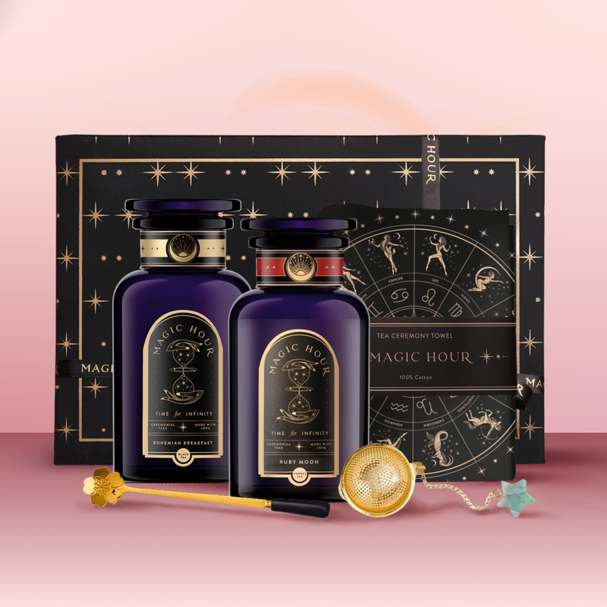 The Wake Up & Wind Down Tea Gift Set features two dark purple "Magic Hour" bottles, a tea ceremony towel, gold tea infuser, golden wand stirrer, and a wrapped crystal arranged before a starry black box—a true tea ritual experience.
