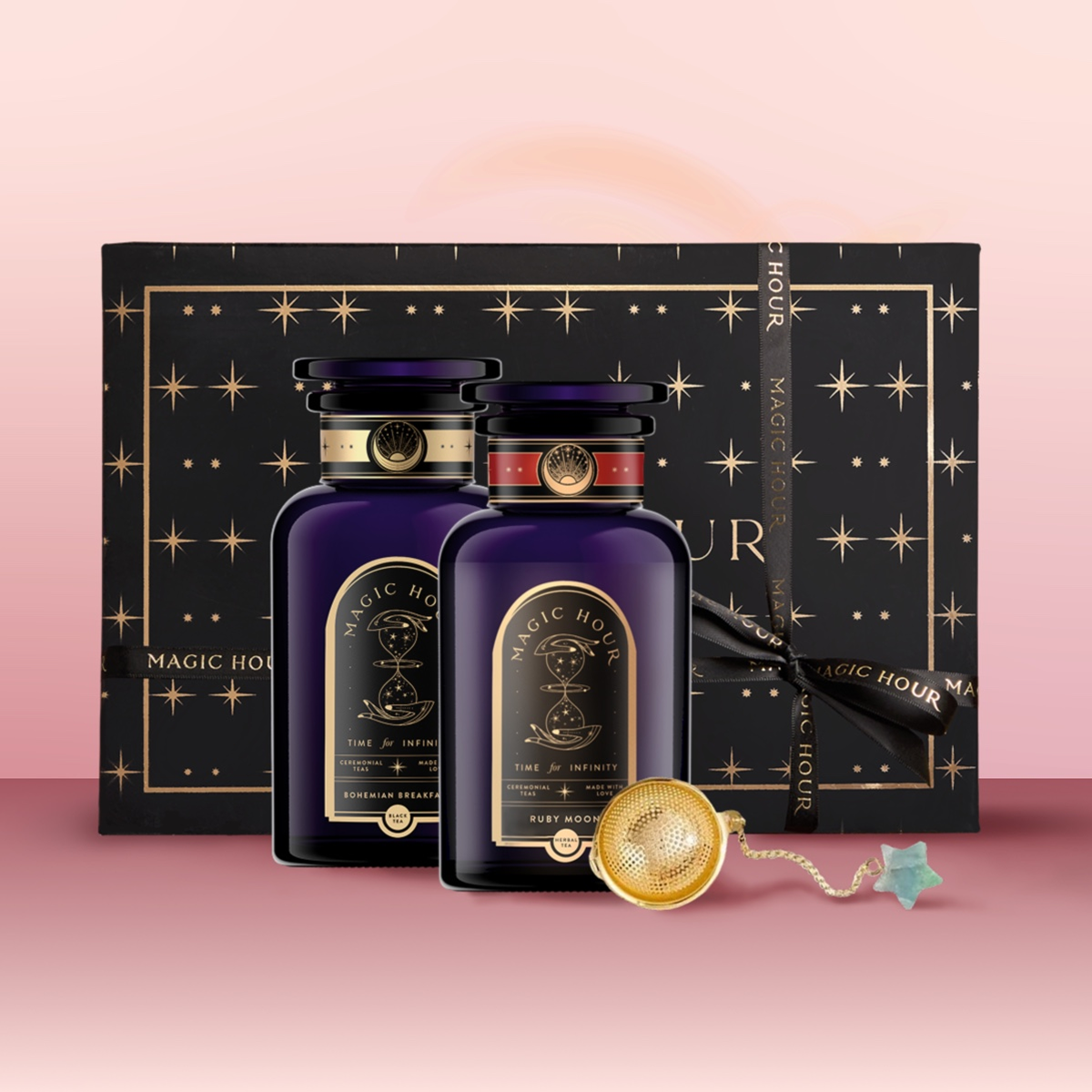 The Wake Up & Wind Down Tea Gift Set features two dark purple and gold “Magic Hour” tea jars, an elegant black gift box with gold starburst designs, a tea infuser, and a star-shaped charm on a pink surface.