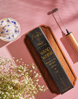 A Wand Whisk: Rechargeable Stainless Steel Eco-Frother, a whisk, a wooden box labeled "Wand Whisk," and a ceramic teacup with white frothy liquid topped with blue flower petals are arranged on a pink surface. Surrounded by white baby's breath flowers, this delicate scene invites you to enjoy the enchantment of Magic Hour Tea.