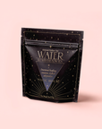 A black, star-patterned pouch of "Water White Tea: Soothing Lemon Balm Rose Reishi Tea for Going with the Flow" by Magic Hour stands upright on a light pink background, featuring gold accents and elegant text on the calming tea blend’s packaging.