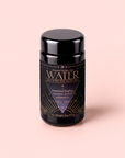 A black cylindrical tea tin labeled “WATER GO WITH THE FLOW TEA” by Magic Hour, adorned with gold and purple accents, rests on a light pink background. This calming herbal blend, Water White Tea: Soothing Lemon Balm Rose Reishi Tea for Going with the Flow, features soothing white tea paired with lemon balm, rose, and reishi mushroom for a tranquil experience.